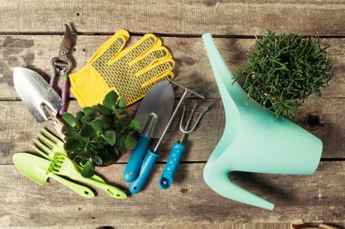 The image displays a collection of gardening tools and supplies arranged on a weathered wooden outdoor surface, likely part of a garden or patio area in Covent Garden. Visible elements include a pair of yellow gardening gloves, a small hand trowel with a metallic blade, a pair of pruning shears with purple handles, a small green hand rake, and a four-pronged cultivator with blue handles. Additionally, there is a light blue plastic watering can and a potted plant with dense, dark green foliage, possibly a herb or small shrub, positioned towards the upper right. The outdoor environment suggests natural daylight, highlighting the textures of the wood, plant leaves, and gardening equipment. This setup reflects typical garden maintenance activities such as planting, watering, pruning, and cultivating, which professionals from Landscapers Covent Garden might assist with to keep outdoor spaces well-maintained and visually appealing, especially in the vibrant outdoor environment of central London.