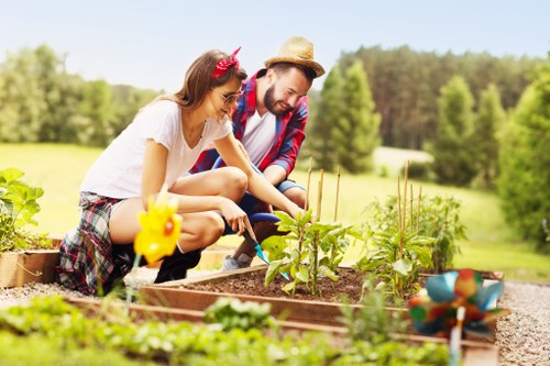 A young girl and a man are working together in a well-maintained garden, planting and tending to vegetable seedlings in raised wooden beds filled with rich soil. The girl, wearing a white t-shirt, plaid shorts, and a pink headband, is actively engaged in planting or watering the plants, while the man, dressed in a plaid shirt and straw hat, is assisting or supervising her activity. The garden features healthy, leafy green plants growing in the beds, with a colorful decorative garden ornament visible in the foreground. The background showcases a lush, green landscape with a variety of mature trees and open grassland under bright, sunny weather, suggesting a peaceful outdoor setting. The scene emphasizes outdoor lawn and garden maintenance, typical of gardening services offered in Covent Garden area, with natural tones and textures of soil, plants, and wood. This depiction underscores residential or community gardening activities supporting local outdoor care, landscaping, and vegetable cultivation efforts.