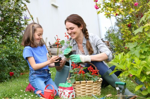 A woman and a young girl are engaging in gardening in an outdoor space, sitting on a lush green lawn with various plants and flowering bushes surrounding them. The woman, wearing green gardening gloves and a light grey shirt, is smiling as she holds a small potted plant with red flowers, while the girl, dressed in a blue t-shirt and denim overalls, gently touches the plant. In front of them, there is a wicker basket, a small red watering can, and a decorated plant pot, suggesting they are planting or tending to flowers. The garden features a mix of vibrant greenery, flowering shrubs, and some larger plants in the background, with a white building partially visible behind the foliage. The scene appears to be set on a mild, sunny day, suitable for outdoor gardening activities, reflecting a typical garden setup that an experienced landscaping service like Landscapers Covent Garden could help develop or maintain as part of outdoor garden care or flower bed design services.