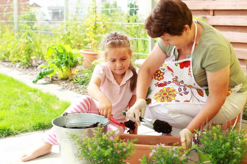 A woman and a young girl are gardening together outdoors in a well-maintained backyard, with a lush vegetable patch and flowering plants visible in the background. The woman, wearing an apron with a floral pattern and gardening gloves, is leaning over a rectangular flower bed filled with dark soil, helping the girl plant or tend to the plants. The girl, dressed in light clothing with pink stripes, is sitting on the paved patio area, actively involved in the gardening activity, holding a small gardening tool. Near them, there is a metal watering can and a terracotta pot, suggesting ongoing planting work. The garden features a neatly trimmed lawn in the foreground, bordered by vibrant green foliage and a wooden fence. The overall scene is bright, indicating a clear day with natural sunlight, and the environment reflects a nurturing outdoor space typical of residential gardens in Covent Garden or nearby London areas, supporting professional gardening services offered by Landscapers Covent Garden.