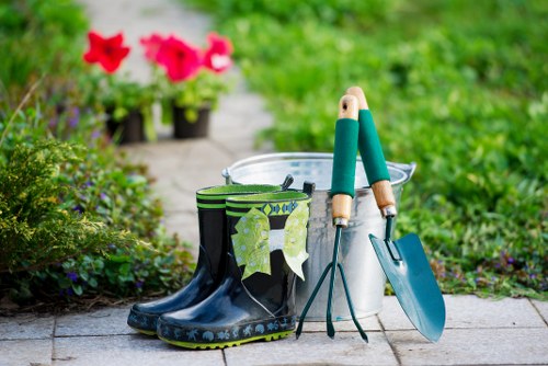 The image shows a garden scene with gardening tools and accessories arranged on a paved pathway. In the foreground, there is a pair of black and green rubber gardening boots placed next to a metal bucket, which contains a trowel and a hand fork with wooden handles, both with green grips. A small spade with a blue handle is leaning against the bucket. Behind these tools, there are two pots of vibrant pink flowers with lush green foliage, adding colour to the scene. The background features a well-maintained garden area with a lawn, dense shrubbery, and flowering plants, suggesting a tidy outdoor space suitable for gardening and landscape maintenance services, especially in the Covent Garden area. The lighting appears to be natural daylight, highlighting the fresh and inviting atmosphere of the garden, indicating a typical outdoor scene prepared for gardening work or outdoor beautification, consistent with services offered by Landscapers Covent Garden.