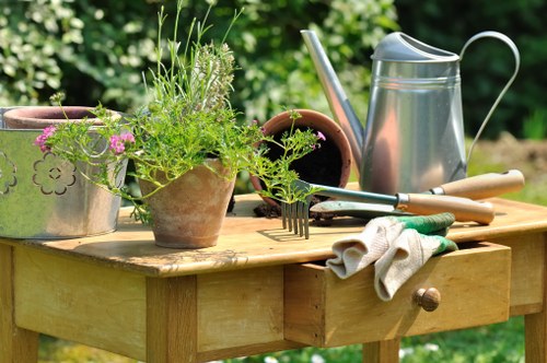 A close-up view of a wooden garden table set outdoors in a natural setting, with a background of lush green foliage. The table holds a variety of gardening tools and accessories, including a silver metal watering can, a pair of pruning shears, a garden fork, and a pair of gardening gloves, which are draped over the edge of the table. Several clay pots are also present, one containing a thriving plant with green, feathery foliage and small pink flowers, while others are empty or contain soil. The scene suggests an early or late sunny day, with natural light illuminating the diverse textures of the wood, metal, clay, and plant surfaces. This outdoor workspace reflects a well-maintained garden environment typical of professional gardening services in Covent Garden, focusing on the preparation and care of flower beds, borders, and outdoor plant arrangements. The overall setting emphasizes readiness for planting or garden maintenance activities, aligned with landscaping and gardening expertise offered by landscaperscoventgarden.co.uk.