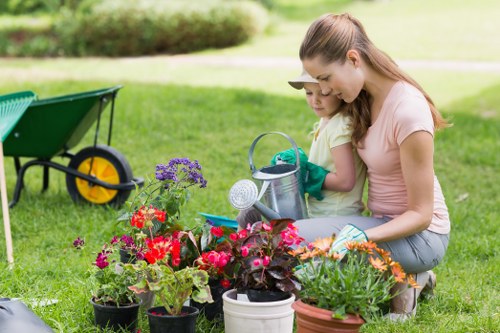 A woman and a young girl are tending to a collection of vibrant flowering plants in a well-maintained garden located in Covent Garden, London. The garden features a lush, green lawn with dense grass, bordered by a variety of potted flowers showing bright red, purple, and orange blooms, and leafy green foliage. In the background, there is a neatly trimmed hedge and a paved patio area. The woman is kneeling on the grass, wearing casual clothing, while the girl stands close beside her, both engaged in gardening activities. Scattered gardening tools such as a watering can and pot are visible, indicating recent planting or maintenance work. The scene is illuminated by natural daylight, suggesting a clear, mild weather day, and captures a peaceful outdoor environment suitable for family gardening or outdoor leisure. This image reflects professional gardening efforts typical of Landscapers Covent Garden, emphasizing garden care, planting, and outdoor beautification services in central London.