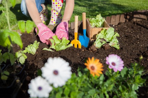 The image shows a garden bed with dark, moist soil, bordered by small bricks, where a child wearing pink gardening gloves is planting or tending to leafy green vegetables, possibly lettuce or similar salad greens. Surrounding the planting area, there are vibrant flowers including white, purple, and orange blooms in the foreground, providing natural colour contrast. The background features neatly maintained grass or lawn, with sunlight illuminating the scene, suggesting a warm and sunny day. Several small hand tools, such as a yellow rake and a blue trowel, are embedded in the soil near the child's hands, indicating active gardening work. The environment appears to be a well-cared-for outdoor space, suitable for gardening and outdoor activities, typical of residential or community gardens in Covent Garden or nearby areas. This scene exemplifies practical gardening activity, involving planting and tending to vegetable beds and decorative flowers, aligning with services offered by landscapers in the region.