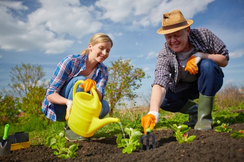 A man and a woman working together in a well-maintained outdoor garden space, actively planting young green vegetable plants into dark, tilled soil. The woman, wearing a blue plaid shirt and orange gardening gloves, is smiling as she holds a yellow watering can, while the man, dressed in a straw hat, grey checked shirt, and green rubber boots, is using a small garden fork to loosen the soil around the plants. Behind them, the garden features a backdrop of lush green grass, a few small trees, and a partly cloudy sky, indicating a bright, sunny day. The scene suggests a focus on gardening and outdoor maintenance, aligning with professional landscaping and garden care services offered by Landscapers Covent Garden, supporting the natural growth and health of garden beds in the local area near Covent Garden, London.
