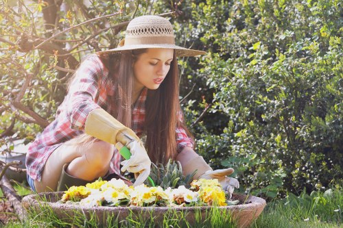 A young woman wearing a wide-brimmed straw hat, plaid shirt, and gardening gloves is kneeling in a landscaped garden with lush greenery and dense shrubs in the background. She is tending to a flower bed bordered by a low stone edge, which contains a variety of blooming flowers in yellow, white, and purple hues. The flower bed is situated on a well-maintained grassy lawn with dense, healthy grass in the foreground, and the garden appears to be part of a residential outdoor space in Covent Garden. The scene is illuminated by natural daylight, suggesting fair weather with a gentle sunlight glow. The overall setting reflects a neat and cared-for garden area, potentially maintained by professional landscapers such as Landscapers Covent Garden, highlighting regular outdoor garden maintenance and flower bed creation services that enhance outdoor aesthetic appeal and natural ambiance.
