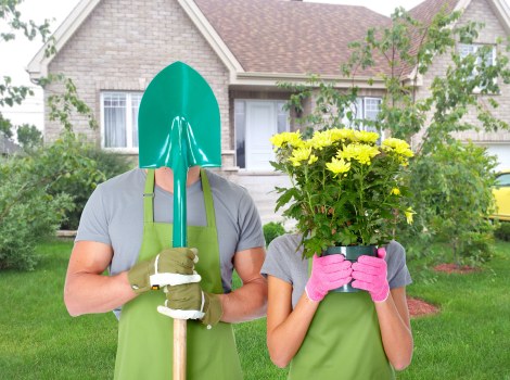 In a residential garden with a well-maintained lawn and flower beds, two individuals wearing green gardening aprons and gloves are standing on the grass. One person is holding a garden shovel with a teal protective cover over their face, and the other is holding a large pink pot filled with blooming yellow flowers, partially obscuring their face. The background features a brick-built house with white-framed windows, a small front porch, and surrounding shrubs and trees, indicating a tidy, landscaped outdoor space typical of gardens in Covent Garden or nearby areas. The scene suggests outdoor gardening or planting activity, supported by gardening services offered by Landscapers Covent Garden, highlighting the lush, healthy grass and vibrant plant life in a peaceful, suburban setting under natural daylight.
