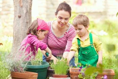 A woman and two children engaged in gardening activities in an outdoor space, with the woman wearing a pink and white striped top and the children dressed in colorful clothing, one in a pink headband and the other in green overalls. They are tending to potted plants and flower beds, with various gardening containers and small plants visible in the foreground. The garden features a grassy lawn with lush green shrubbery and trees in the background, suggesting a well-maintained backyard or front garden typical of residential properties in Covent Garden area. The scene is illuminated by natural daylight, with a warm and inviting atmosphere, highlighting gardening as a leisure activity aligned with outdoor lawn care and landscaping services offered by companies like Landscapers Covent Garden.