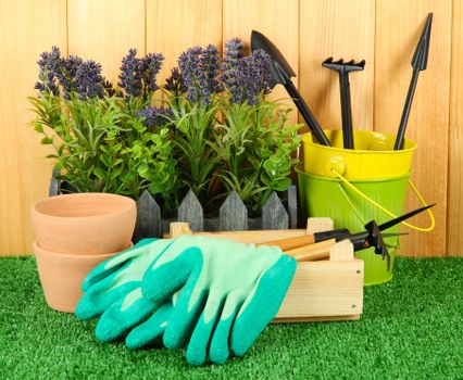 The image depicts a neatly arranged outdoor gardening workspace with a backdrop of a warm-toned wooden fence. In the foreground, there is a patch of artificial green grass serving as a surface for gardening tools and supplies. Prominently featured are several terracotta plant pots stacked in front of purple-flowering lavender plants, which are well-maintained and healthy, situated behind a row of small, dark grey garden borders. To the right, a bright yellow plastic bucket holds a variety of garden tools including a hand rake, trowel, and pruners. In front of the bucket, a pair of turquoise gardening gloves rests on a light wooden tray or small table, alongside a small black hand clipper. The scene reflects an organized outdoor space prepared for planting or garden maintenance, likely part of a gardening service in Covent Garden, suitable for enhancing flower beds, shrubbery, or curb appeal in a city garden setting. The overall environment appears to be well-lit, suggesting daytime with natural sunlight illuminating the various garden elements.